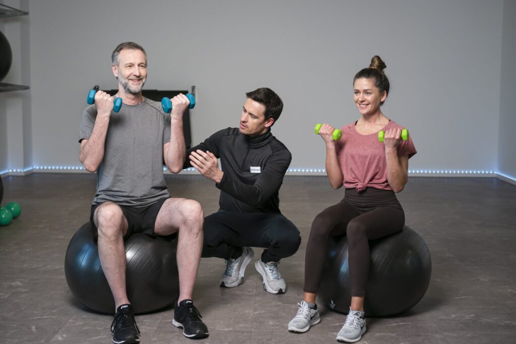 Zwei Personen führen mit Kurzhanteln eine Übung im Sitzen auf Gymnastikbällen durch, betreut von einem Sporttherapeuten