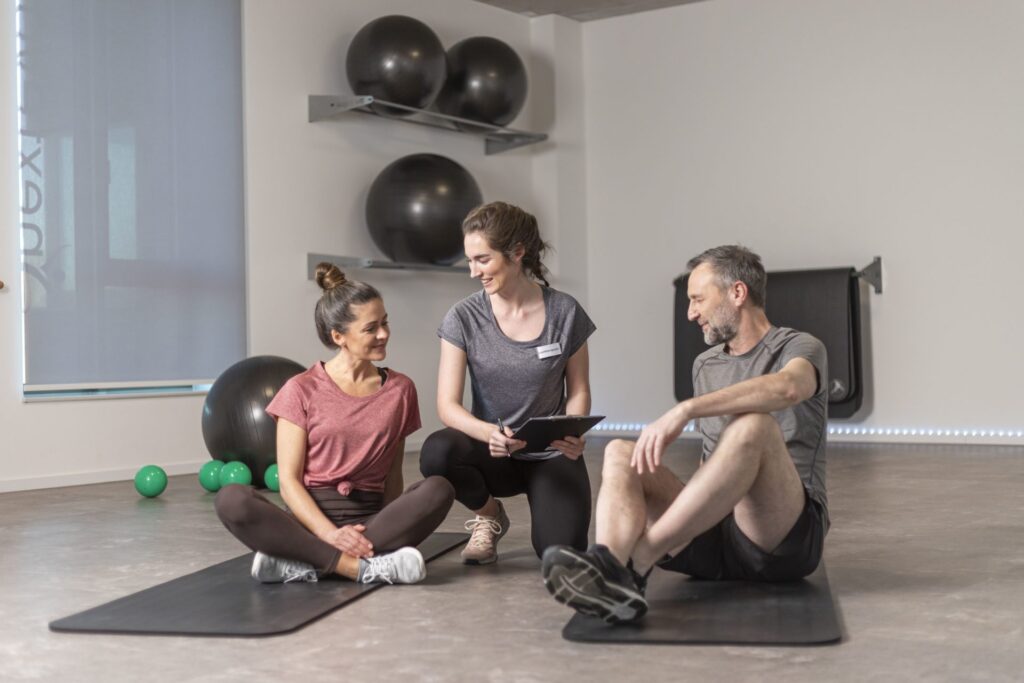 Zwei Personen sitzen in Sportkleidung auf Matten, eine Therapeutin mit Klemmbrett spricht freundlich mit ihnen