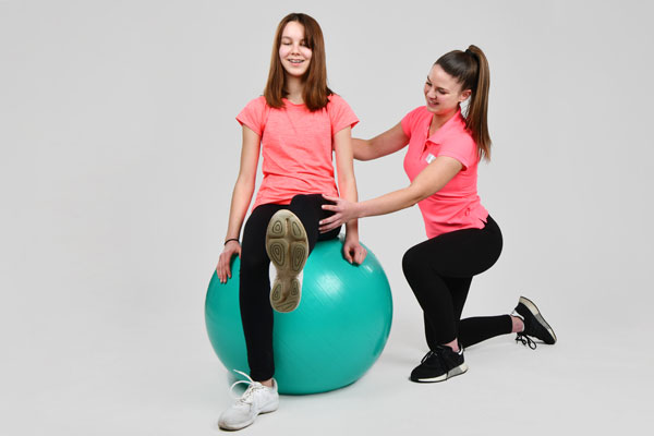 Ein Mädchen sitzt auf einem Gymnastikball und hebt ein Bein, während eine Therapeutin sie dabei unterstützt