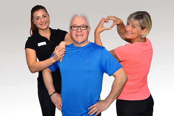 Älterer Mann in blauer Sportkleidung mit zwei Frauen – eine Therapeutin und eine Frau formt ein Herz mit den Händen
