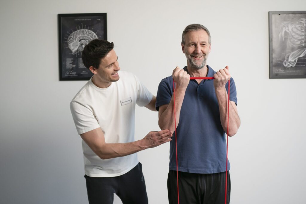 Ein Physiotherapeut begleitet einen älteren Mann bei einer Übung mit rotem Theraband im Stehen